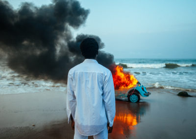 Kojo stares at a burning car on a beach.